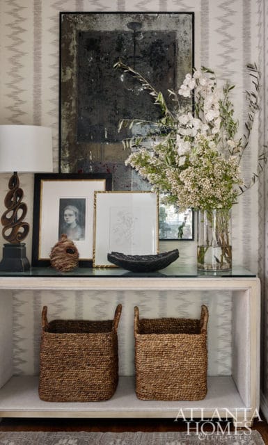 Console table with a glass top displaying framed artwork, a vase of white flowers, and decorative objects. Two woven baskets sit on the shelf below.