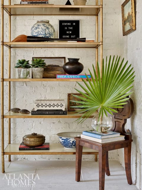 Gold etagere filled with decorative objects including vases, books, plants, and a framed painting against a white brick wall.