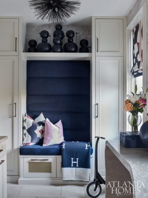 Built-in banquette with navy blue tufted upholstery, geometric and watercolor pillows, and a navy blanket with white 'H' logos. Above, a shelf displays black abstract sculptures and a spiky light fixture.