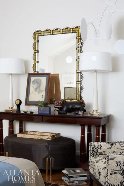 Dark wood console table topped with a framed portrait, a black carved animal sculpture, and two white lamps. A dark upholstered ottoman sits below the table.