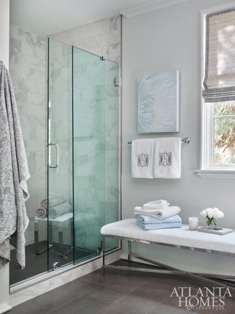Modern bathroom with glass-enclosed shower featuring marble tile, a white upholstered bench with stacked towels, and a window with a roman shade.