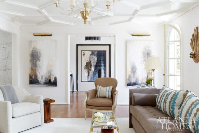 Living room with white walls, coffered ceiling, abstract art, tufted sofa, leather armchair, and glass coffee table.