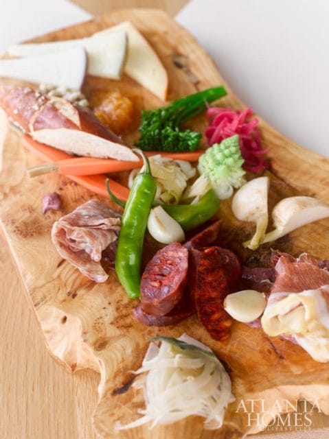 Charcuterie board with sliced meats, cheeses, pretzel, peppers, garlic, and pickled vegetables on a live-edge wooden board.