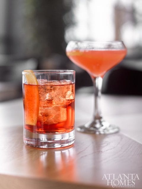 Two red cocktails with ice and garnishes on a wooden table, with the Atlanta Homes & Lifestyles logo in the lower right corner.