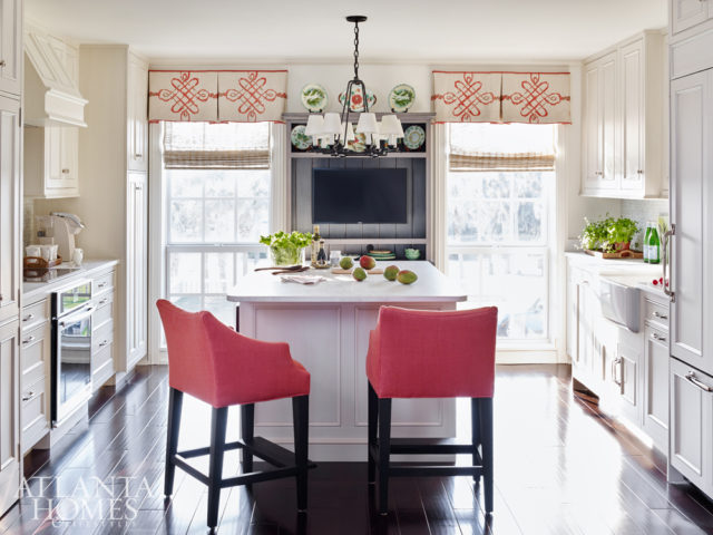 Watermelon red adds oomph to a pair of linen-covered counter stools in the kitchen, which was completely redone with the help of a local contractor. The sophisticated scroll pattern on the valances comes from an Osborne & Little fabric, beautifully flanking a small collection of the homeowners’ own colorful pottery.