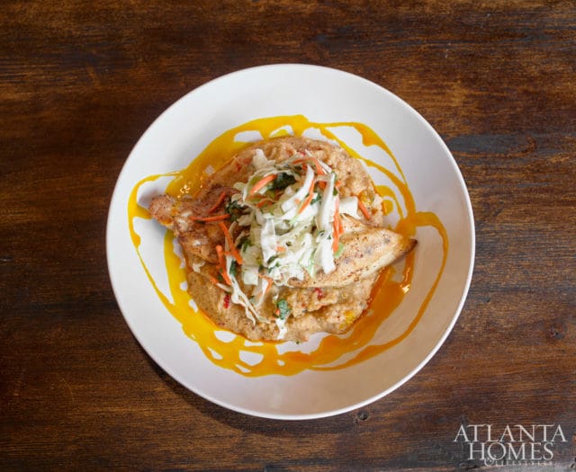 White bowl of pan-seared fish with grits and coleslaw, drizzled with orange sauce, on a dark wood table. Atlanta Homes & Lifestyles logo in the bottom right corner.