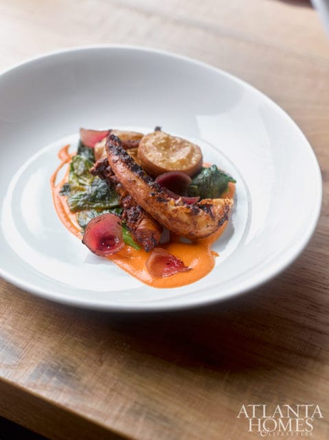 Grilled octopus tentacles served with roasted potatoes, greens, and a vibrant orange sauce on a white plate.