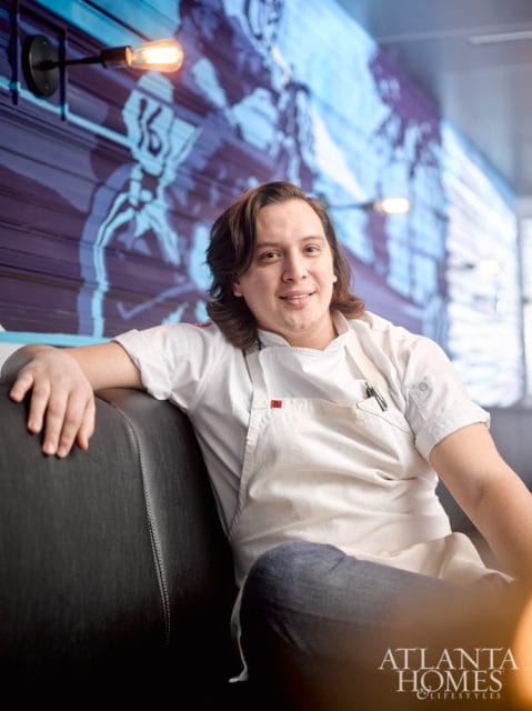 Chef in white chef coat and apron sitting on a dark leather couch in front of a blue mural and industrial lights.