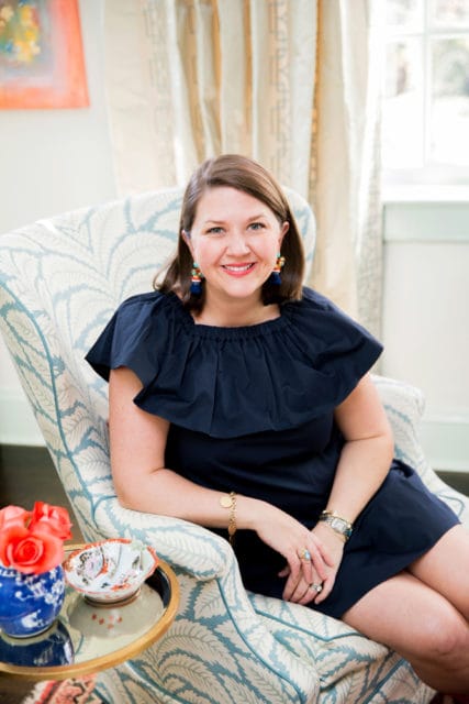 Woman with brown hair wearing a navy blue dress and colorful earrings, sitting in a patterned armchair.