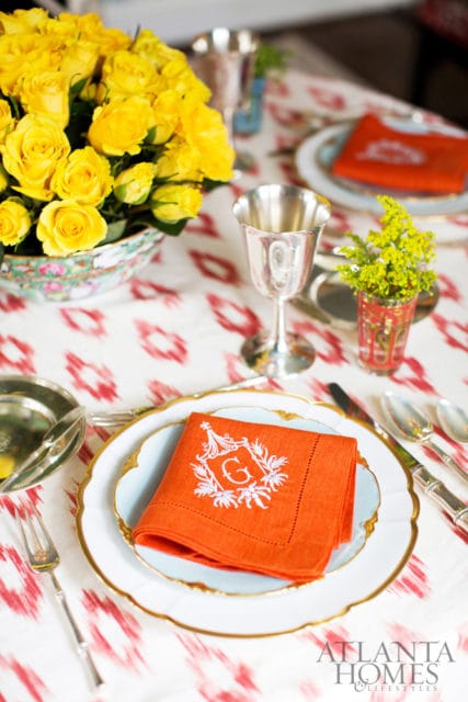 Orange linen napkin with white embroidered monogram 'G' in a decorative crest, folded on a light blue plate with gold trim.