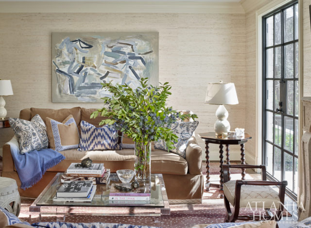 Grasscloth walls warm up the cozy family room, where the homeowners’ sofas, upholstered in a Sunbrella velvet, were updated with an elegant tape trim and accented by a glass-and-Lucite coffee table, abstract artwork by Brian Coleman and a nearly indestructible antelope-pattern rug by Stark. The ceramic gourd lamps are the homeowners’ own. Brass fruits, speckled glass bowl and pottery on side table, Townhouse by Robert Brown.