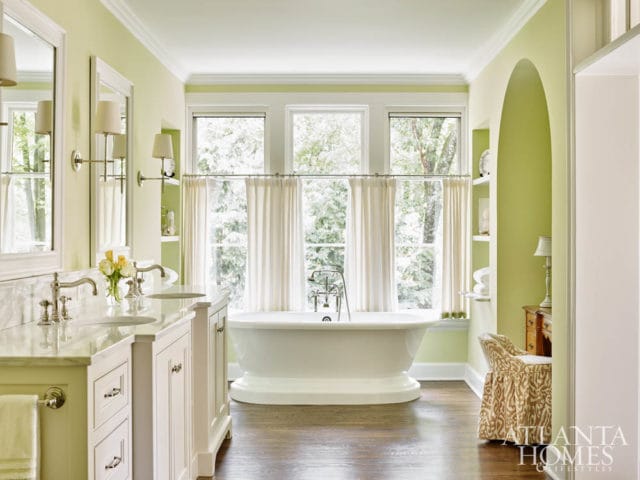 Light green bathroom with white double vanity, marble countertops, freestanding bathtub, and a patterned upholstered chair.