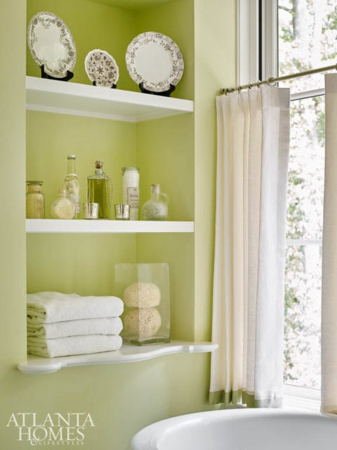 Pale green bathroom with white shelves displaying decorative plates, bottles, jars, and stacked towels. A glass vase holds two natural sponges. White curtains hang beside a window.
