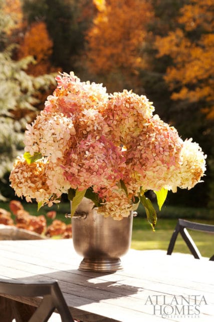 A bouquet of pink and white hydrangeas in a silver bucket sits on a wooden table, with autumn foliage in the background.