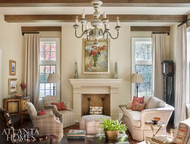 Living room with a stone fireplace, plush seating, and exposed wooden beams on the ceiling. A painting of flowers hangs above the fireplace.