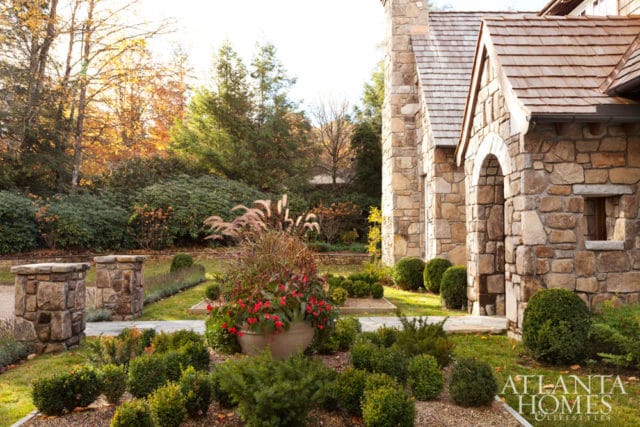 Stone house with arched entryway and cedar shake roof, surrounded by manicured boxwood hedges and ornamental grasses in autumn