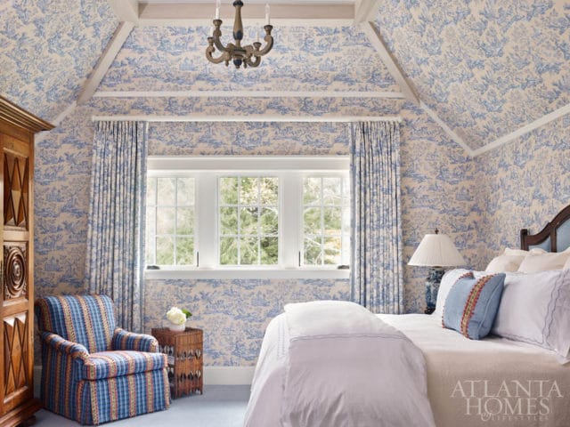 Bedroom with blue and white toile wallpaper on walls and ceiling, white trim, wooden armoire, plaid armchair, wicker side table, and a bed with white linens and blue pillows.