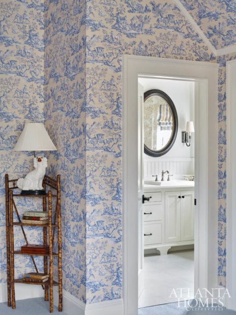 Hallway with blue and white toile wallpaper, a bamboo etagere with books and a white dog statue, and a view into a white bathroom with a round mirror.
