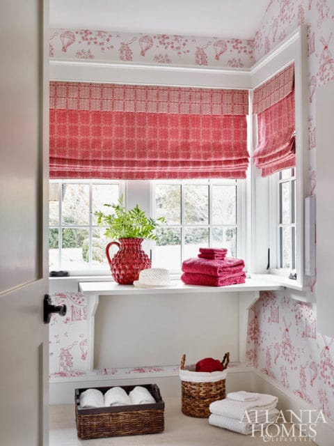 Corner of a room with red and white toile wallpaper, red patterned Roman shades, a white shelf with a red vase, folded towels, and a woven basket with towels.