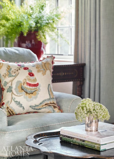 Light blue armchair with a patterned throw pillow, a silver vase of white flowers, and books on a dark tray. A large red vase with greenery sits behind the chair.