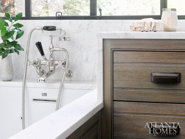 White bathtub with marble tile backsplash and a vintage-style chrome faucet with black handles. Adjacent is a dark wood vanity with fluted drawers and bronze hardware.