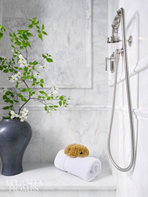 Marble tiled bathroom with a dark gray vase of white dogwood branches, a rolled white towel with a natural sponge, and a chrome showerhead.