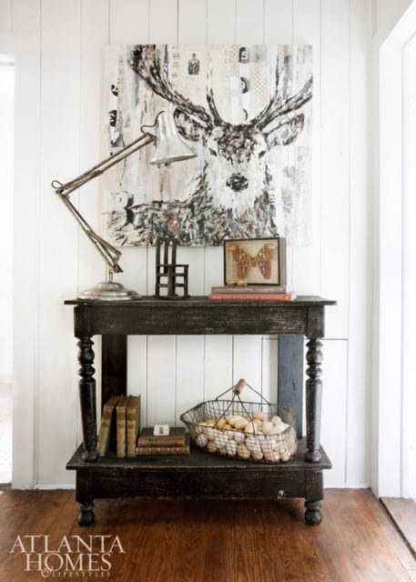 Black distressed console table with a metal desk lamp, abstract deer artwork, and a wire basket filled with eggs.