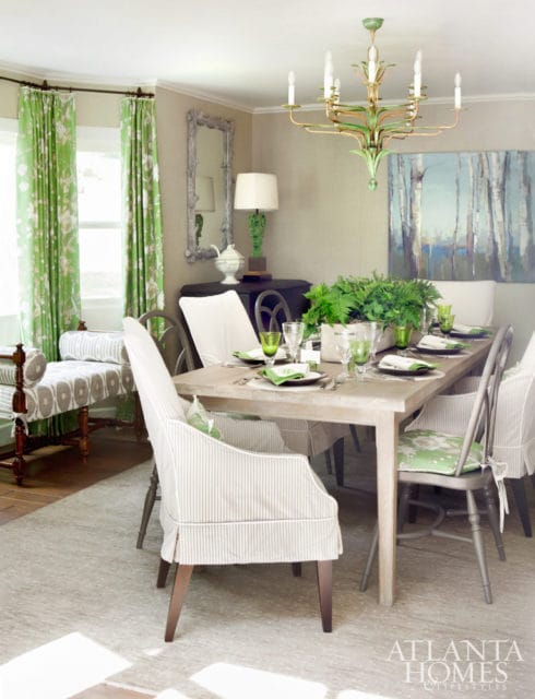 Dining room with a light wood table set for a meal, upholstered chairs with striped slipcovers, green floral curtains, and a green chandelier.