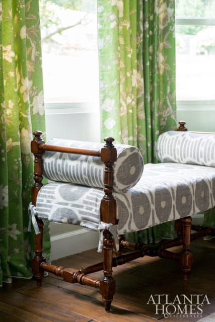 Antique wooden bench with grey and white patterned upholstery, set against bright green floral curtains.