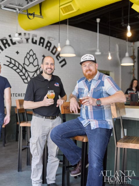 Two men stand and sit at a bar in a brewery with a "BarleyGarden" logo on the wall behind them. The man on the left wears a black polo shirt and khaki pants, holding a beer. The man on the right wears a blue plaid shirt and jeans, also holding a beer.