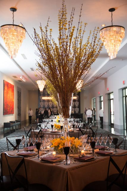 Elegant dining setup in a prefunction room with tables set with tan linens, dark glassware, and tall floral arrangements of yellow flowers and branches. Crystal chandeliers hang from the ceiling.