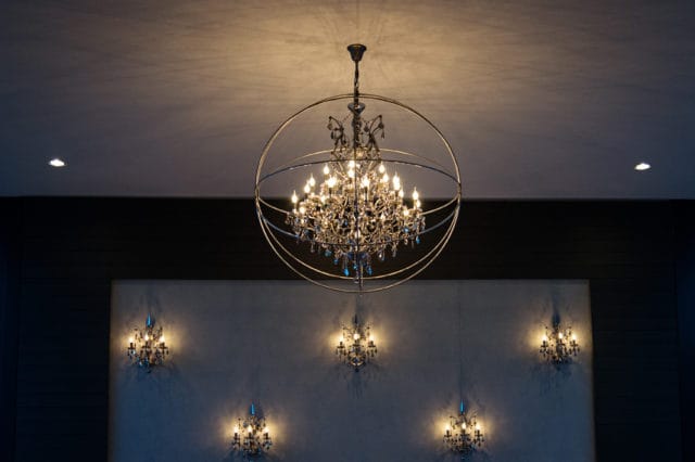 A grand chandelier with multiple lights and crystal accents hangs from the ceiling within a large circular metal frame. Below, five smaller wall sconces with similar crystal details illuminate a textured wall.