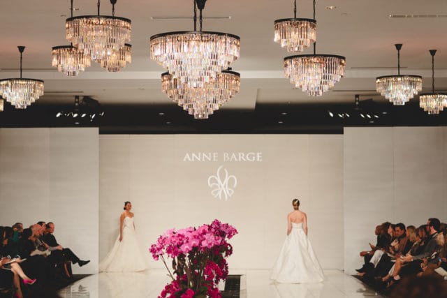 Wedding dress runway show with models in white gowns, audience seated on the sides, and ornate crystal chandeliers overhead.