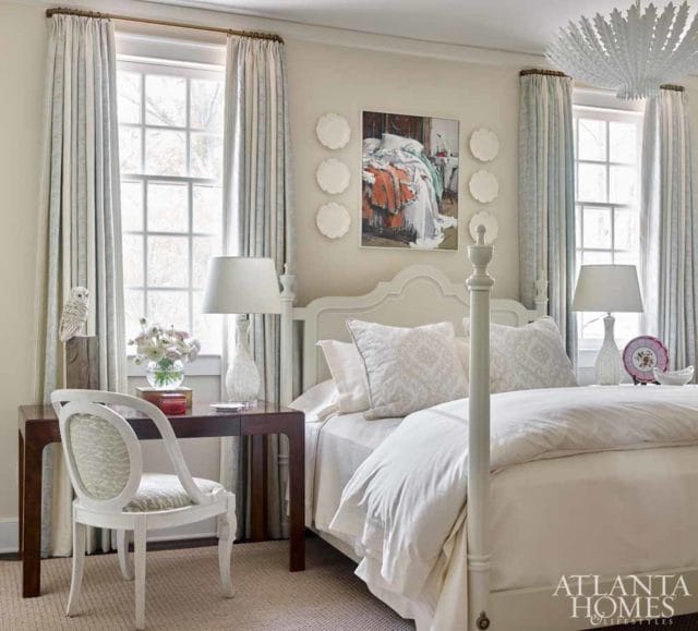 Bedroom with a white four-poster bed, patterned curtains, and a dark wood desk with a white chair. A painting hangs above the bed.