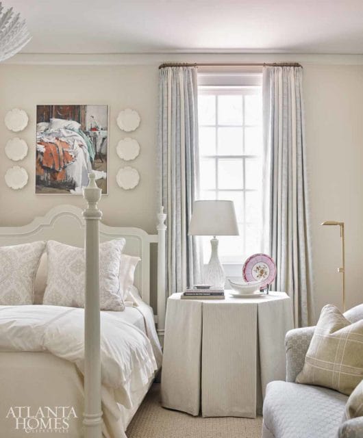 Bedroom with a white four-poster bed, patterned pillows, and a painting of a rumpled bed on the wall. Light blue and white patterned curtains hang over a window.