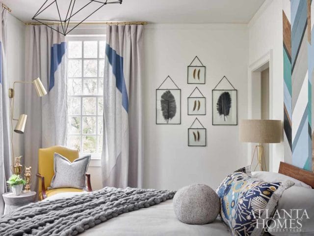 Bedroom with gray chunky knit blanket, yellow armchair, and wall art featuring framed feathers. A patterned pillow on the bed has the Atlanta Homes & Lifestyles logo.