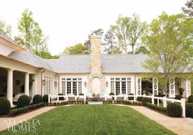 White house with gray roof, stone chimney, and white fence surrounding manicured lawn and topiaries.