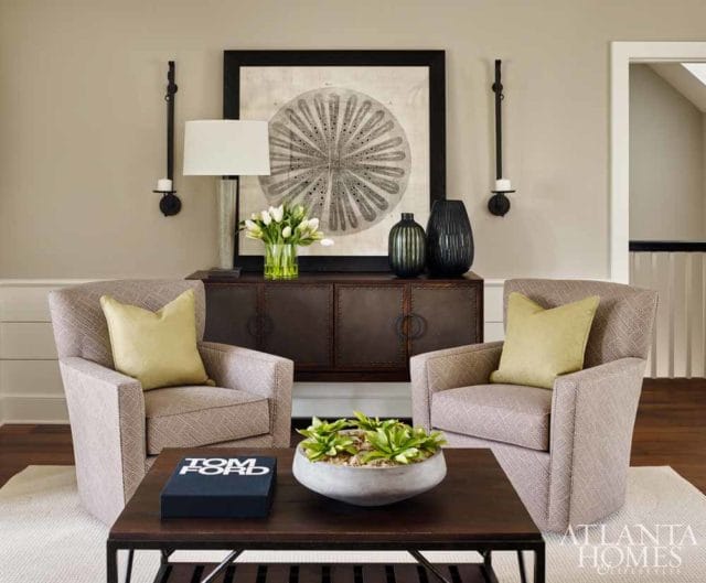 Two upholstered armchairs flank a dark wood console table with a large framed artwork and vases. A coffee table with a book and plant sits in front.