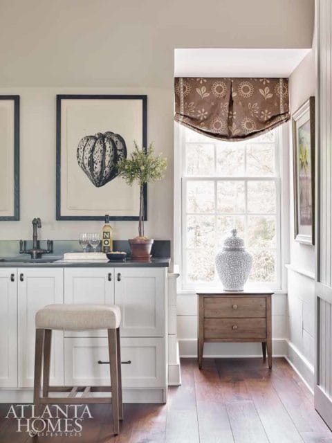 White kitchen cabinetry with dark hardware, a framed botanical print, and a neutral upholstered stool. A window with a floral Roman shade is visible.