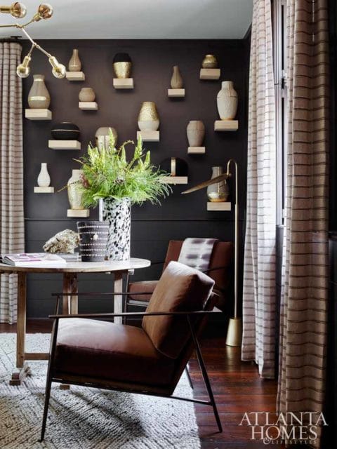 Dark gray room with a wall of shelves displaying various decorative vases and vessels, a round wooden table with chairs, and a floor lamp.
