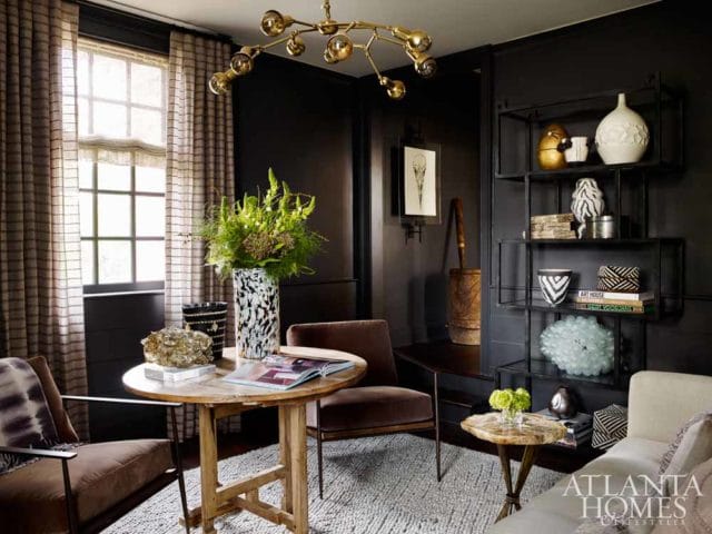 Dark room with a round wooden table, two chairs, and a shelf unit displaying various decorative objects. A large vase with greenery sits on the table.