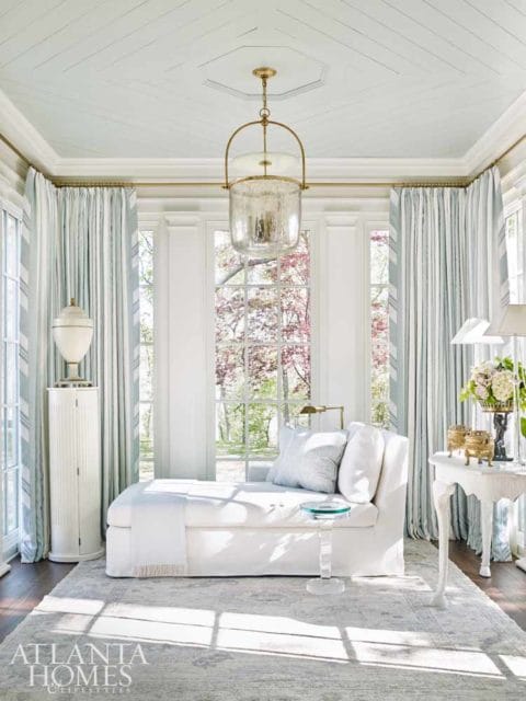 Sunlit room with a white chaise lounge, pale blue striped curtains, a decorative white cabinet, and a large glass pendant light hanging from a coffered ceiling.