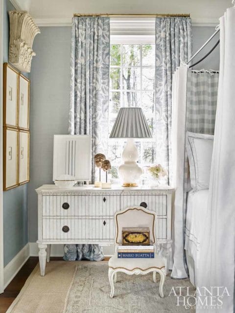 Bedroom with light blue walls, a striped dresser, a canopy bed with white linens, and a window with patterned curtains.