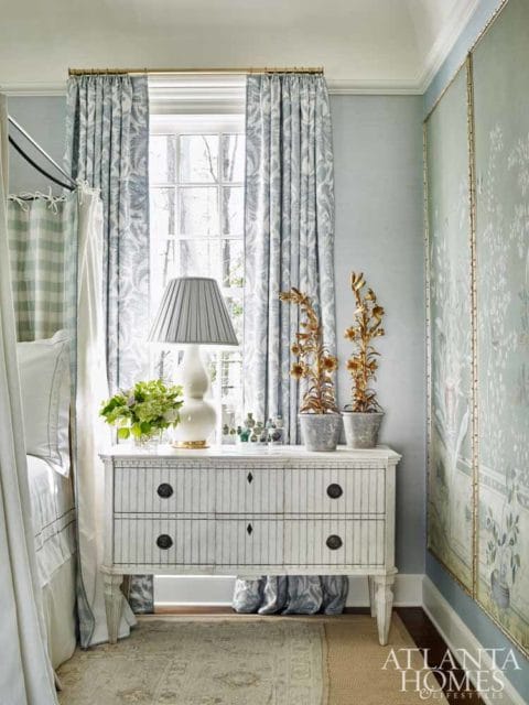 Bedroom with a white dresser, blue floral curtains, and a large chinoiserie wall panel. A white lamp sits on the dresser next to two potted plants.