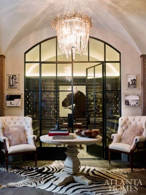 Elegant interior with two tufted armchairs, a round pedestal table with books and a wooden bowl, a zebra print rug, and a glass-enclosed wine cellar with a horse head sculpture.