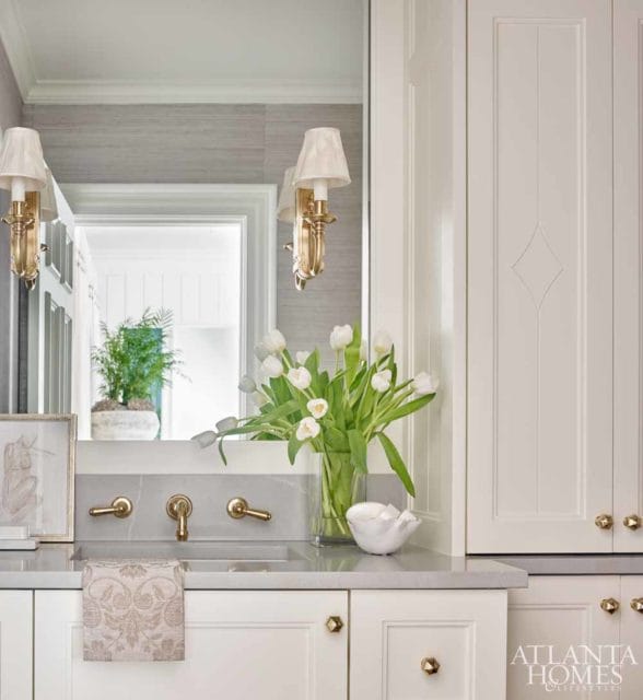 Cream cabinetry with brass hardware, gray stone countertop, and a sink with brass faucets. A vase of white tulips sits on the counter, reflected in the mirror above.