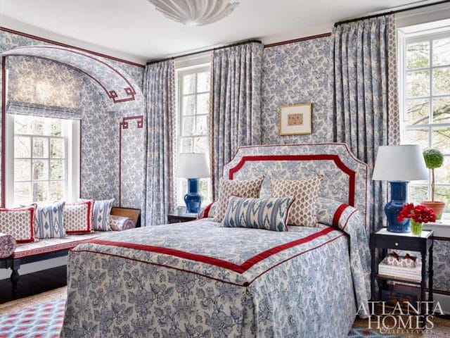 Blue and white floral wallpaper and bedding in a bedroom with a red-trimmed headboard, blue lamps, and a window seat.