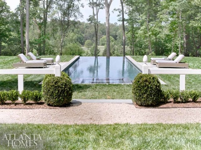 Infinity pool with lounge chairs on deck, surrounded by manicured lawns, lush trees, and a distant view of rolling hills.