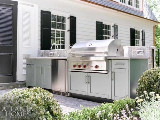 Outdoor kitchen with stainless steel grill, sink, and light gray cabinetry set against a white house with black shutters.