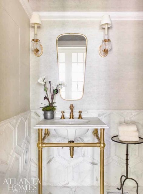 Powder room with marble hexagon tile wainscoting, gold-framed mirror, brass faucet, and gold pedestal sink base.